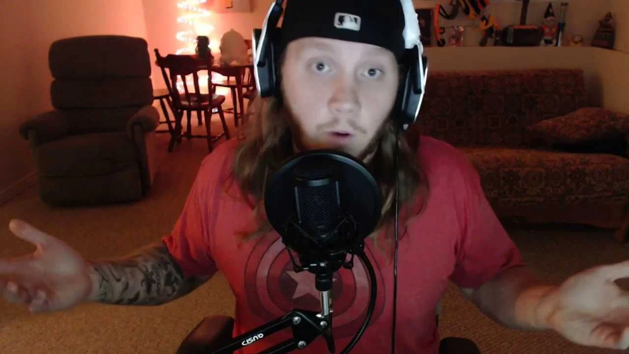 TimTheTatman wearing headphones and a backward cap, speaking into a microphone with his arms spread.